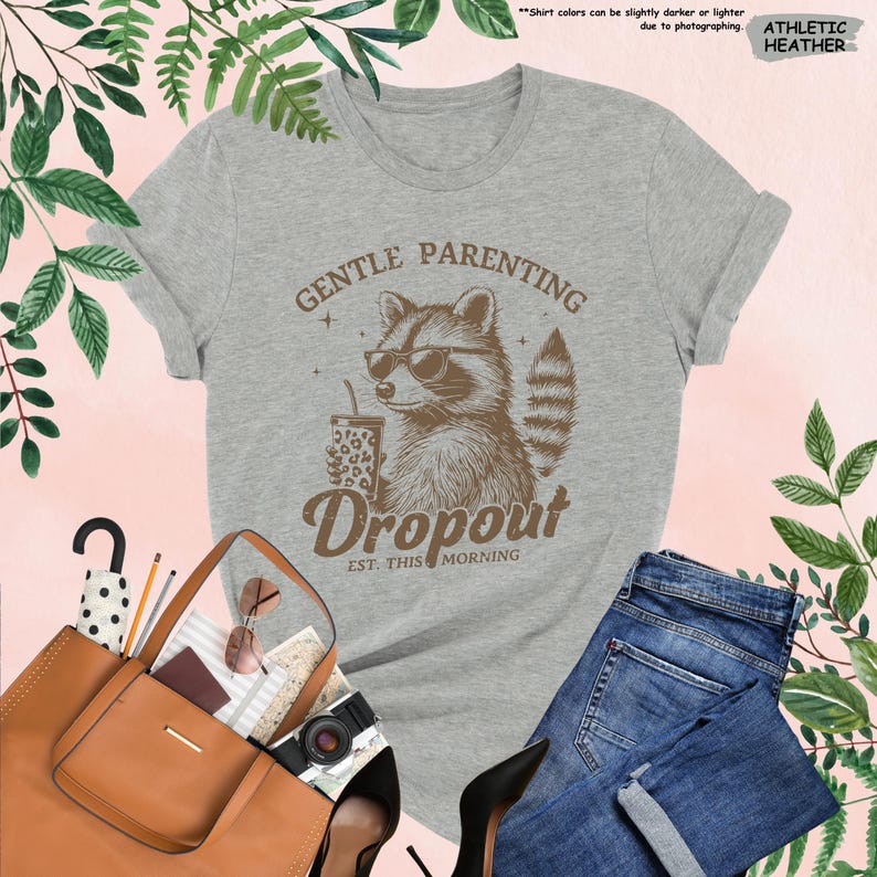 May include: Athletic heather gray t-shirt with a raccoon wearing sunglasses and holding a drink. The shirt has the words "GENTLE PARENTING Dropout EST. THIS MORNING" printed on it. The shirt is paired with blue jeans, a brown tote bag, and black heels.