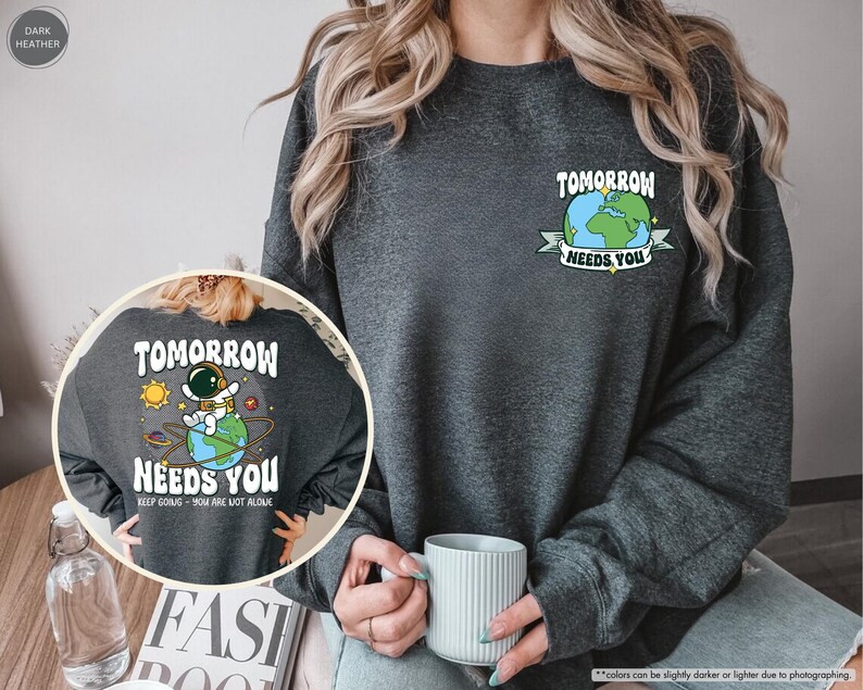May include: Dark grey sweatshirt featuring the text "TOMORROW NEEDS YOU" and an astronaut graphic. The design includes a cartoon astronaut on Earth with the text "KEEP GOING - YOU ARE NOT ALONE". A smaller version of the design is on the upper right.