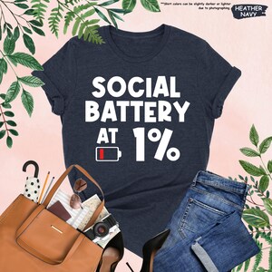 May include: Heather navy t-shirt with the text "SOCIAL BATTERY AT 1%" in white. The design includes a battery icon at 1%. The shirt is paired with blue jeans, a brown tote bag, and black heels.