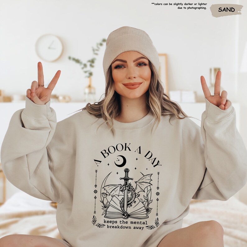 a woman sitting on a bed wearing a book a day sweatshirt