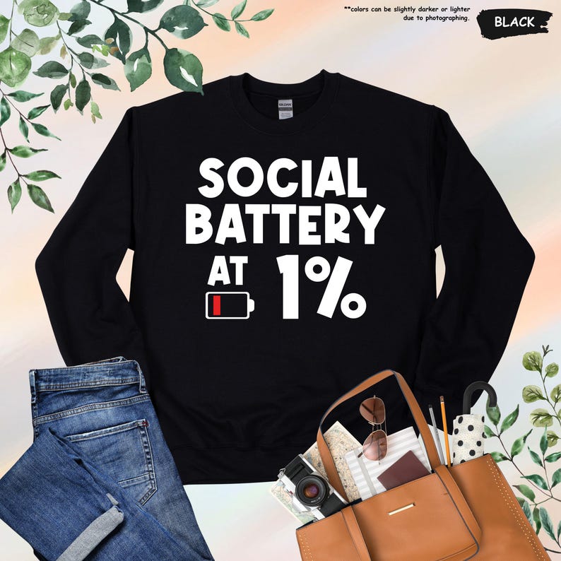May include: Black sweatshirt with the text "SOCIAL BATTERY AT 1%" in white. The design includes a battery icon with a low charge indicator. The sweatshirt is displayed with a pair of blue jeans and a brown tote bag.