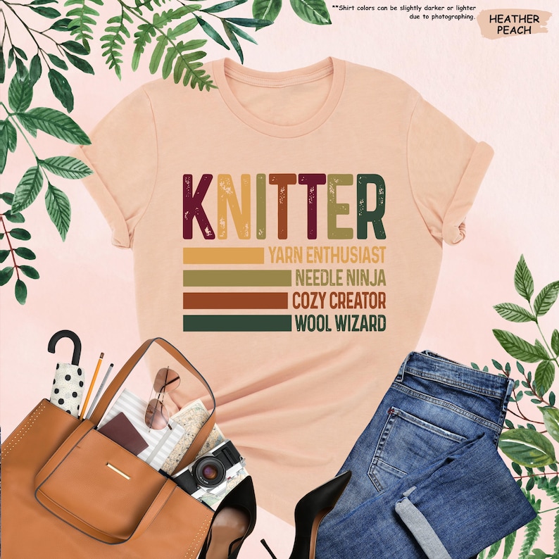 May include: A heather peach t-shirt with the text "KNITTER" in a vintage style font. The text is layered over a series of colorful stripes with the words "Yarn Enthusiast", "Needle Ninja", "Cozy Creator", and "Wool Wizard" written below.