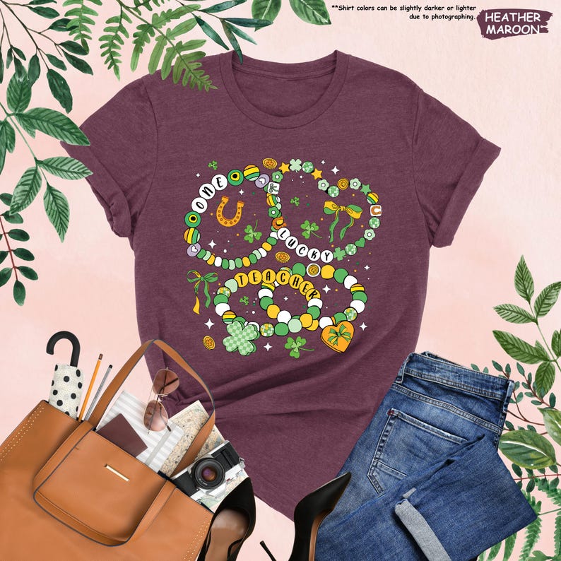 K&ouml;nnte beinhalten: Ein T-Shirt in Heide-Maroon mit einem Motiv aus gr&uuml;nen, gelben und goldenen Kleeblatt-Halsketten mit den Worten "One Lucky Teacher" darauf.