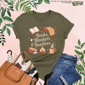May include: Heather olive green t-shirt with the text "Books, Blankets & Bonfires" in a whimsical font. The graphic includes illustrations of a book, blanket, bonfire, and a mug. The shirt is paired with blue jeans and a brown tote bag.