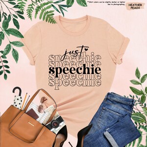 Puede incluir: Una camiseta de color melocot&oacute;n con el texto "just speechie" en letras negras repetidas en un patr&oacute;n apilado.