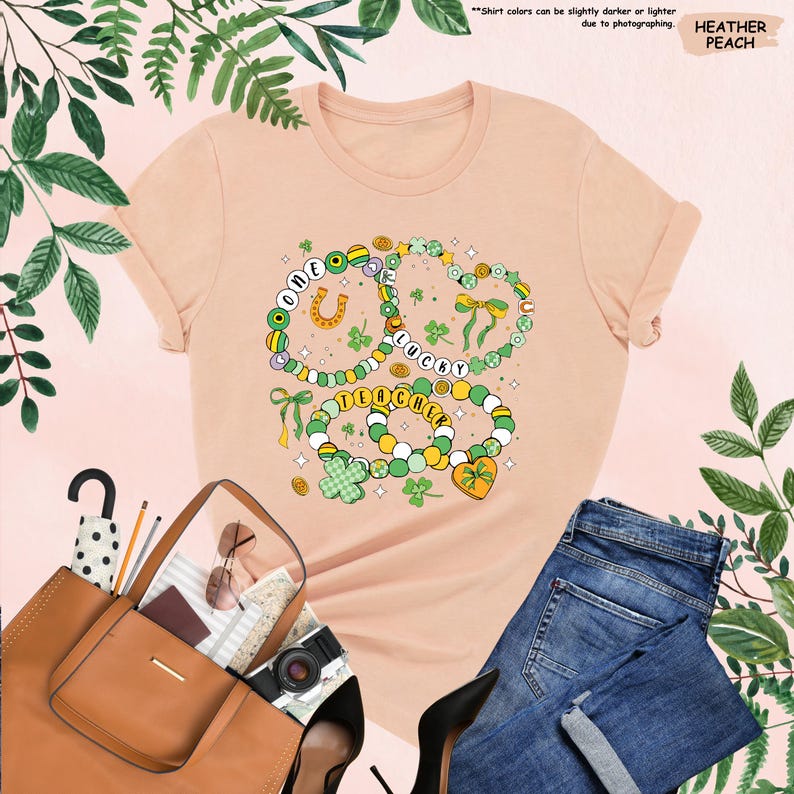 K&ouml;nnte beinhalten: Ein pfirsichfarbenes T-Shirt mit einem Motiv aus drei Halsketten mit Anh&auml;ngern. Die Halsketten haben gr&uuml;ne, gelbe und wei&szlig;e Perlen mit goldenen Akzenten. Die Anh&auml;nger umfassen ein Hufeisen, ein Herz und eine Schleife. Der Text auf den Halsketten lautet "One Lucky Teacher".