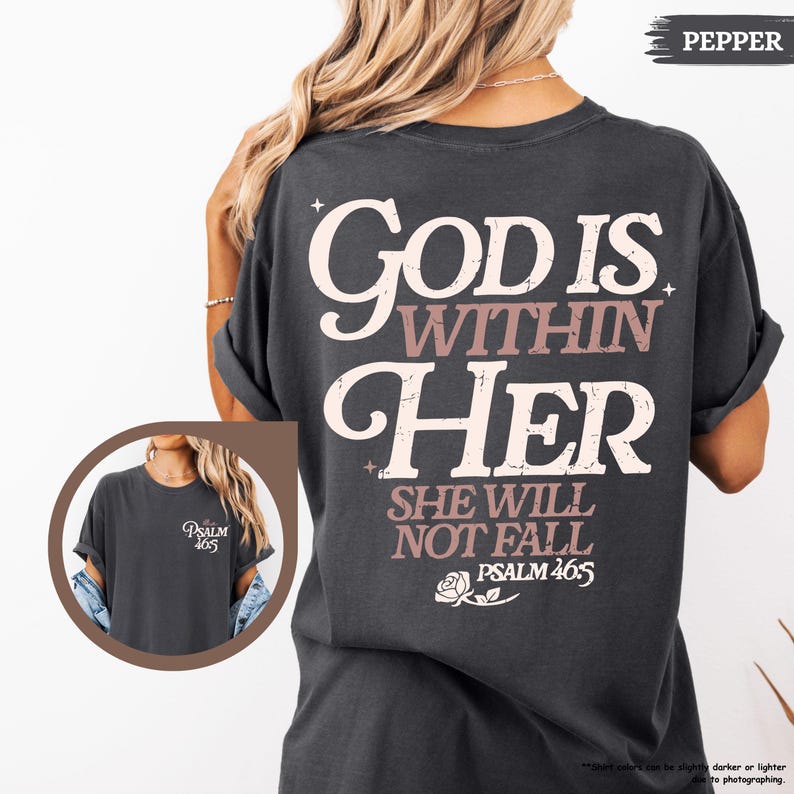 May include: Dark gray t-shirt with the text "God Is Within Her" in white and mauve lettering. The back of the shirt also features the text "She Will Not Fall" and "Psalm 46:5". A small graphic of a rose is below the text.