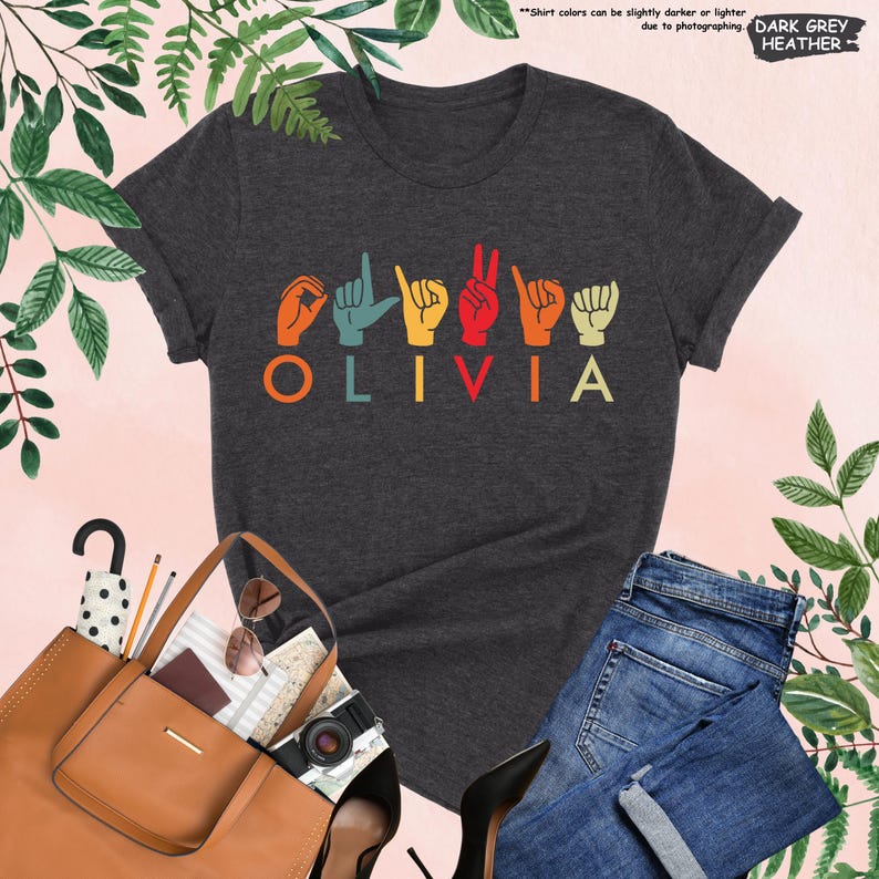 Puede incluir: Una camiseta de color gris oscuro jaspeado con el nombre "OLIVIA" escrito en colores arco&iacute;ris. Las letras est&aacute;n en Lengua de Se&ntilde;as Espa&ntilde;ola.