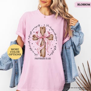 Op de afbeelding: Roze T-shirt met een houten kruis en een roze strik. Het kruis heeft de tekst "Zij lacht zonder angst voor de toekomst" en "Spreuken 31:25".