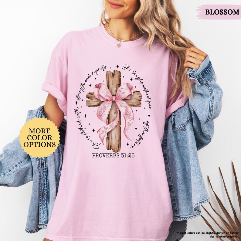 Op de afbeelding: Roze T-shirt met een houten kruis en een roze strik. Het kruis heeft de tekst "Zij lacht zonder angst voor de toekomst" en "Spreuken 31:25".