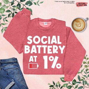 May include: Heather scarlet red sweatshirt with the text "SOCIAL BATTERY AT 1%" in white. The design includes a low battery icon. A cup of coffee and a pair of blue jeans are also in the image.