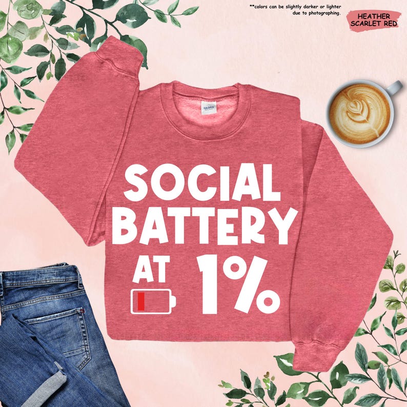 May include: Heather scarlet red sweatshirt with the text "SOCIAL BATTERY AT 1%" in white. The design includes a low battery icon. A cup of coffee and a pair of blue jeans are also in the image.