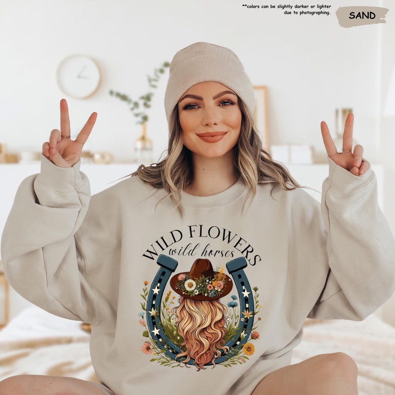 May include: A light beige sweatshirt with a graphic design of a woman with long blonde hair wearing a brown cowboy hat with flowers. The graphic also includes a blue horseshoe with stars and flowers. The text "WILD FLOWERS wild horses" is printed on the sweatshirt.