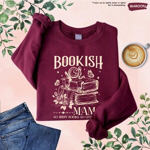 May include: A maroon sweatshirt with the text "BOOKISH MAM" in a vintage style. The design includes a stack of books, a clock, butterflies, and roses. The phrase "SO MANY BOOKS SO LITT" is also present. A pair of blue jeans and a cup of coffee are also in the image.