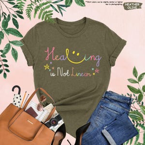 K&ouml;nnte beinhalten: Ein gr&uuml;nes T-Shirt aus Heather-Material mit dem Text "Healing is Not Linear" in bunten Buchstaben mit einem Smiley und Sternen.