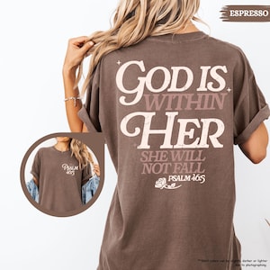 May include: Espresso-colored t-shirt with the text "God is Within Her" on the back, along with "She Will Not Fall" and "Psalm 46:5." A smaller version of the text "Psalm 46:5" is on the front. The shirt has rolled-up sleeves.