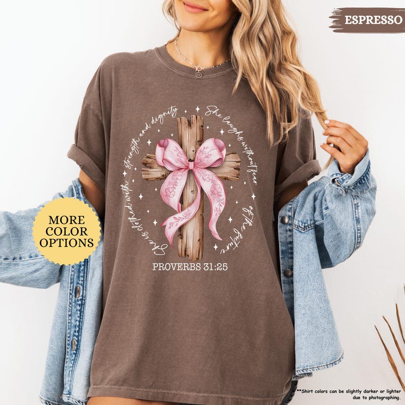 Op de afbeelding: Een bruin T-shirt met een roze lint om een houten kruis. De tekst "She laughs without fear of the future" en "Proverbs 31:25" staat op het T-shirt gedrukt.