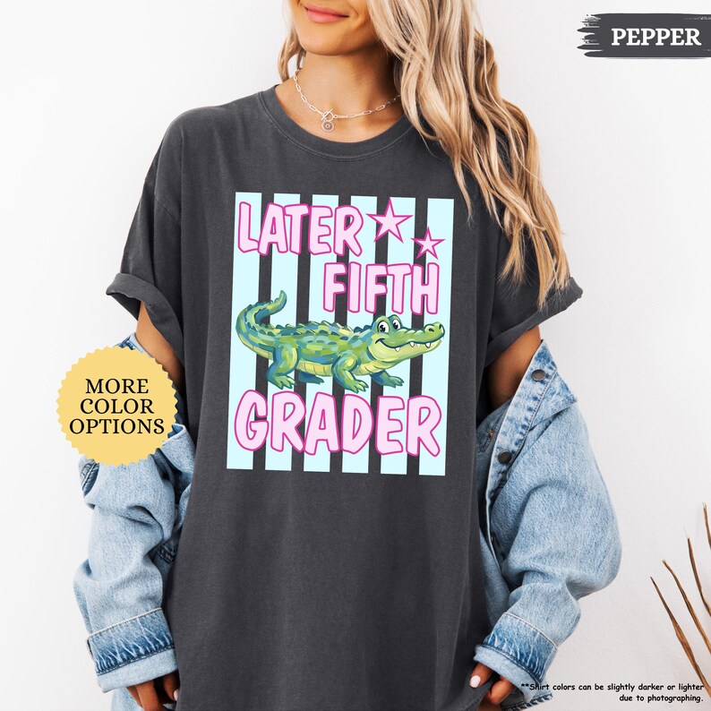 May include: Dark gray t-shirt with a graphic that says "Later Fifth Grader" in pink and white, featuring a cartoon alligator. The shirt has rolled-up sleeves and is worn with a denim jacket.