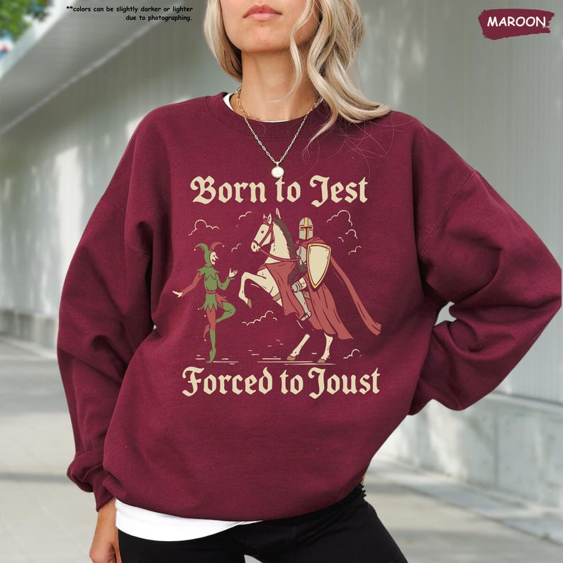K&ouml;nnte beinhalten: Burgunderfarbenes Sweatshirt mit dem Text "Born to Jest, Forced to Joust" und einer Grafik eines Narren und eines Ritters zu Pferd. Das Sweatshirt hat eine einfarbige burgunderrote Farbe.