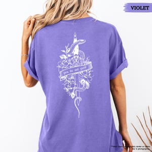 May include: A violet t-shirt with a white graphic on the back. The graphic includes a snake, sword, flowers, and the text "you do not yield". The shirt has rolled-up sleeves.