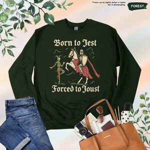 K&ouml;nnte beinhalten: Dunkelgr&uuml;nes Sweatshirt mit dem Text "Born to Jest Forced to Joust" und einer Grafik eines Narren und eines Ritters zu Pferd. Das Sweatshirt wird mit einer blauen Jeans und einer braunen Tragetasche gezeigt.