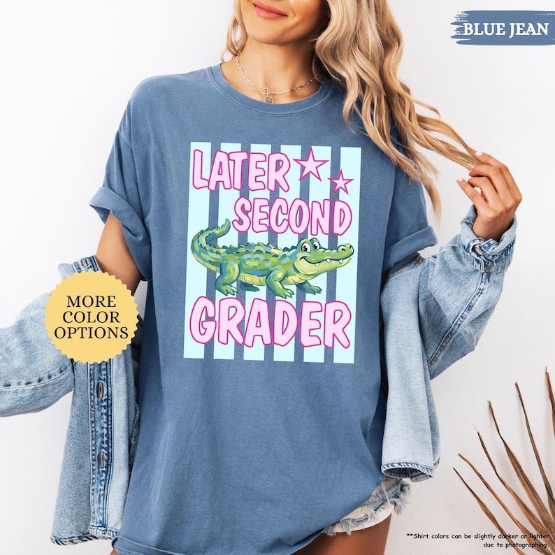 May include: Blue jean-colored t-shirt with the text "Later Second Grader" in pink, featuring a cartoon alligator. The shirt has rolled-up sleeves and a relaxed fit. Additional text on the shirt includes "More Color Options" and "Blue Jean".
