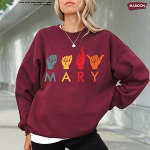 Puede incluir: Una sudadera color granate con el nombre "MARY" escrito en letras de colores. Cada letra est&aacute; representada por un signo de lenguaje de se&ntilde;as.