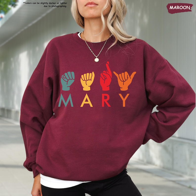 Puede incluir: Una sudadera color granate con el nombre "MARY" escrito en letras de colores. Cada letra est&aacute; representada por un signo de lenguaje de se&ntilde;as.
