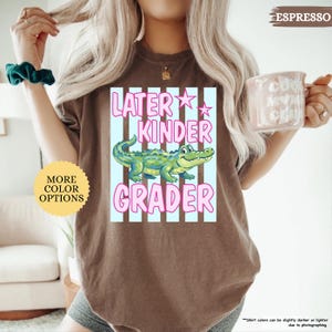 May include: Espresso-colored t-shirt with a graphic that reads "Later Kinder Grader" in pink and white. The graphic features a cartoon alligator in green and blue. A round sticker on the left says "More Color Options".