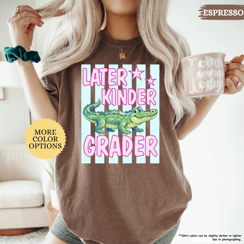 May include: Espresso-colored t-shirt with a graphic that reads "Later Kinder Grader" in pink and white. The graphic features a cartoon alligator in green and blue. A round sticker on the left says "More Color Options".