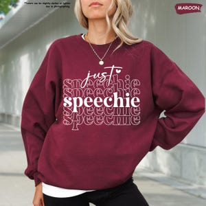 Puede incluir: Una sudadera color granate con el texto "just speechie" impreso en blanco. El texto se repite en un patr&oacute;n apilado con la palabra "speechie" en un tama&ntilde;o de fuente m&aacute;s grande.
