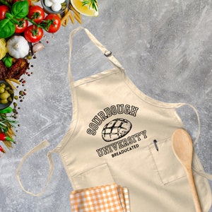 May include: A beige kitchen apron with a black graphic of a sourdough bread loaf and the text "Sourdough University Breadeducated".