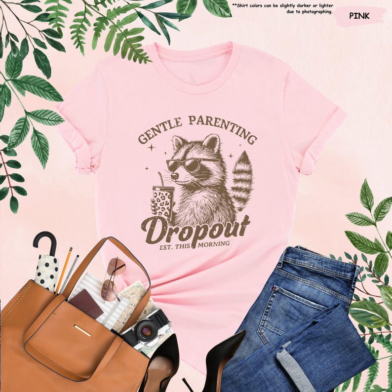 May include: A light pink t-shirt featuring a raccoon wearing sunglasses and holding a drink. The text on the shirt reads "GENTLE PARENTING Dropout EST. THIS MORNING". The shirt is paired with blue jeans, a brown tote bag, and black heels.