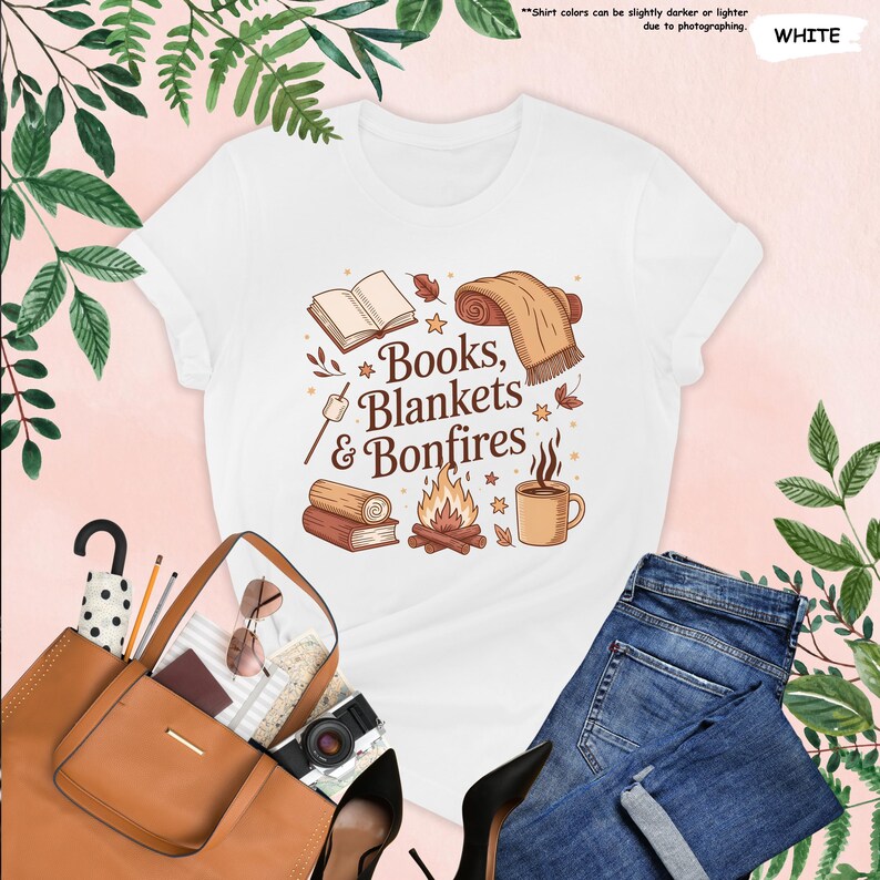 May include: White t-shirt with a graphic design featuring the text "Books, Blankets & Bonfires" with illustrations of a book, blanket, bonfire, and coffee mug. The shirt is paired with blue jeans and a brown tote bag.