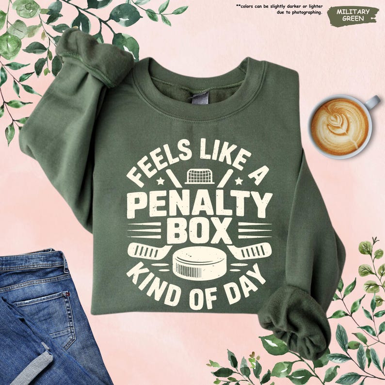 May include: Military green sweatshirt with the text "Feels Like A Penalty Box Kind Of Day" in white. The design includes hockey sticks, a hockey puck, and a hockey net. The sweatshirt is folded on a pink surface.