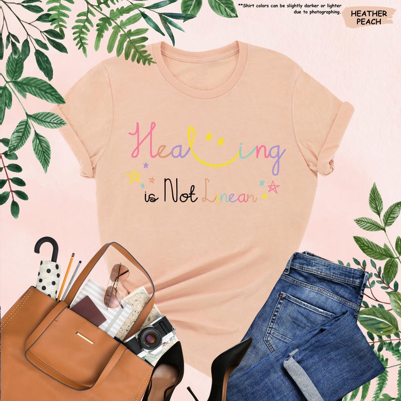 K&ouml;nnte beinhalten: Ein pfirsichfarbenes T-Shirt mit dem Text "Healing is Not Linear" in Regenbogenfarben. Der Text ist in einem verspielten, handschriftlichen Stil mit einem Smiley im Wort "Healing" geschrieben.