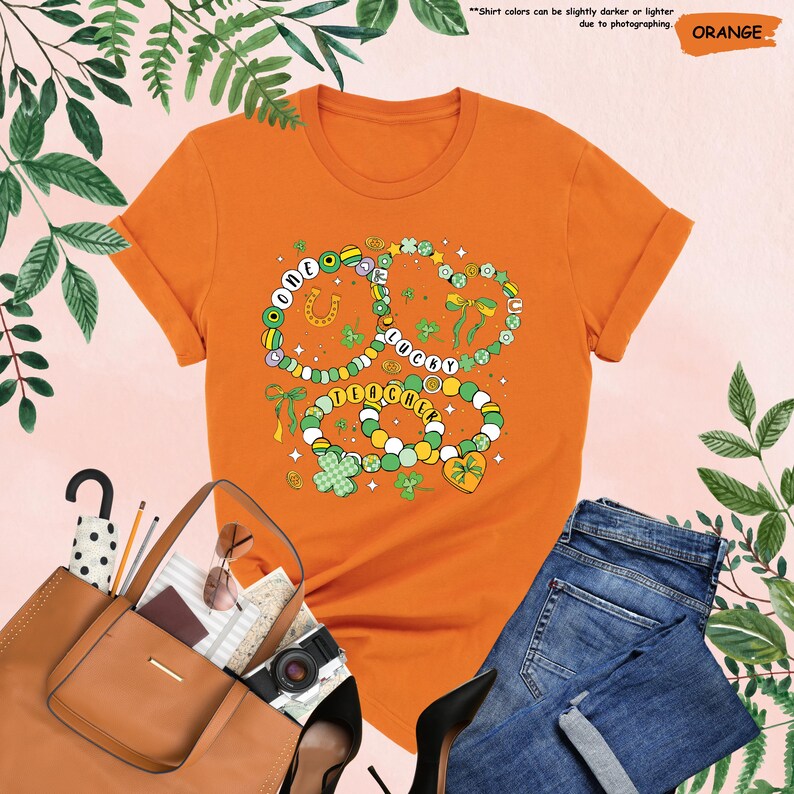 K&ouml;nnte beinhalten: Orangefarbenes T-Shirt mit einem Motiv aus drei Halsketten mit Kleebl&auml;ttern, einem Hufeisen und den Worten "One Lucky Teacher".