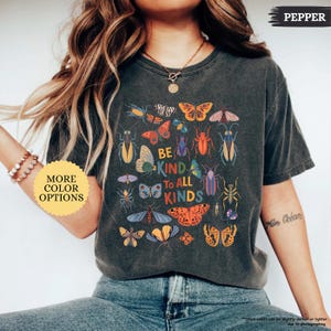 May include: Dark grey t-shirt featuring a colourful insect and butterfly graphic with the text "BE KIND TO ALL KINDS". A yellow circle with the text "MORE COLOR OPTIONS" is in the image.