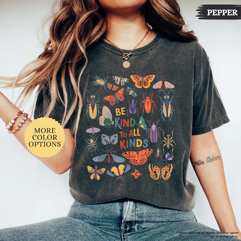 May include: Dark grey t-shirt featuring a colourful insect and butterfly graphic with the text "BE KIND TO ALL KINDS". A yellow circle with the text "MORE COLOR OPTIONS" is in the image.