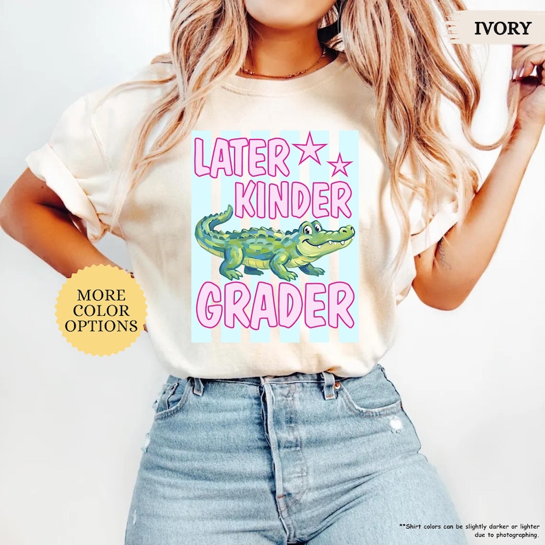 May include: Ivory t-shirt with the text "LATER KINDER GRADER" in pink and a cartoon alligator. The shirt also has a light blue background with vertical stripes and the words "MORE COLOR OPTIONS" in a yellow circle.