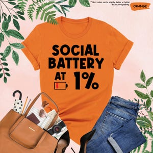 May include: Orange t-shirt with the text "SOCIAL BATTERY AT 1%" in black. The shirt is paired with blue jeans, a brown purse, and black heels. The image includes a camera, sunglasses, and other accessories.