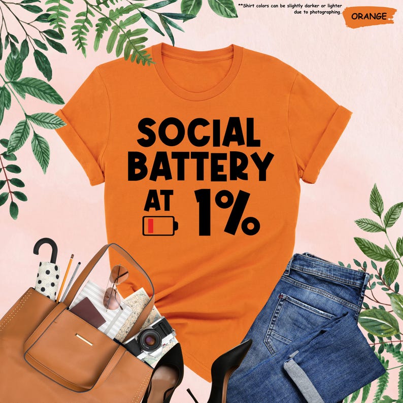 May include: Orange t-shirt with the text "SOCIAL BATTERY AT 1%" in black. The shirt is paired with blue jeans, a brown purse, and black heels. The image includes a camera, sunglasses, and other accessories.