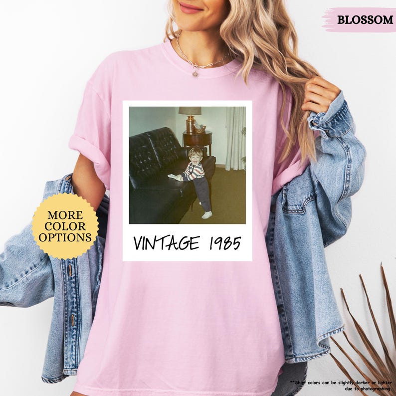 Op de afbeelding: Roze T-shirt met een vintage Polaroid-foto van een klein kind dat op een bank zit. Onder de foto staat de tekst "Vintage 1985".