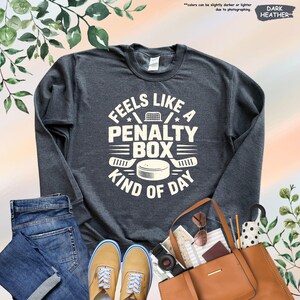 May include: Dark heather gray sweatshirt with the text "Feels Like A Penalty Box Kind Of Day" in white. The design includes a hockey puck, hockey sticks, and a net. The sweatshirt is paired with blue jeans and tan shoes.