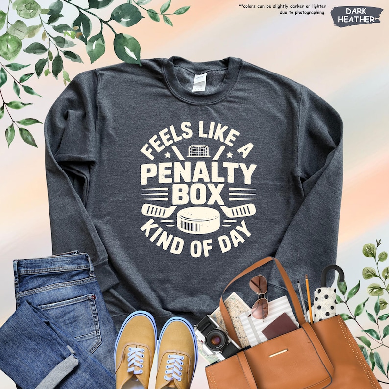 May include: Dark heather gray sweatshirt with the text "Feels Like A Penalty Box Kind Of Day" in white. The design includes a hockey puck, hockey sticks, and a net. The sweatshirt is paired with blue jeans and tan shoes.