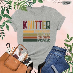 May include: A gray t-shirt with the word "Knitter" in a vintage style font. The text is layered with colorful stripes and the words "Yarn Enthusiast", "Needle Ninja", "Cozy Creator", and "Wool Wizard" are written below.