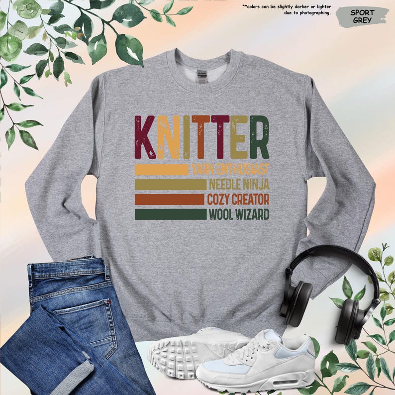 May include: A heather grey sweatshirt with the word "KNITTER" in a distressed, colorful font. Below the word are the phrases "NEEDLE NINJA", "COZY CREATOR", and "WOOL WIZARD". The sweatshirt is paired with blue jeans, white sneakers, and black headphones.