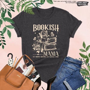May include: Dark grey heather t-shirt with the words "BOOKISH MAMA" and "SO MANY BOOKS SO LITTLE TIME" printed on the front. The design features a stack of books, a clock, butterflies, and flowers. A brown tote bag and blue jeans are also visible.