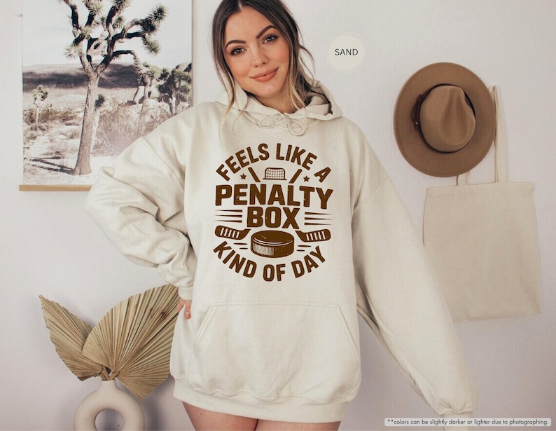 May include: A sand-colored hoodie with a front pocket and a drawstring hood. The front of the hoodie features the text "FEELS LIKE A PENALTY BOX KIND OF DAY" in brown, along with hockey-themed graphics.