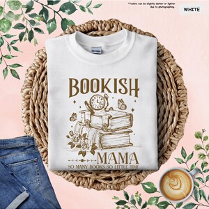 May include: White sweatshirt with the text "BOOKISH MAMA" and a graphic of books, a clock, and butterflies. The phrase "SO MANY BOOKS SO LITTLE TIME" is below the graphic. The sweatshirt is on a woven basket.
